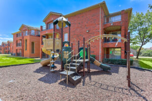 Exterior Playground, Monkey Bars, slides, wood chip soft ground, residential red brick buildings in background, meticulous landscaping, lush foliage, photo taken on a sunny day.